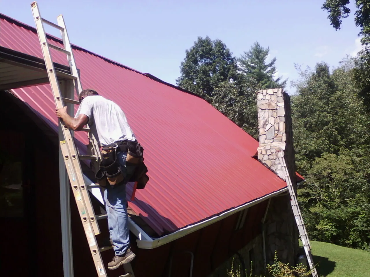 Roofer on a ladder beside a red metal roof and stone chimney — Soffit & Fascia Repair by Roofing Brothers in Madrid